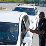 A police officer standing beside a white car reviews a driver’s license handed to him by a woman during a traffic stop. The officer’s patrol vehicle with flashing lights is parked behind the car.