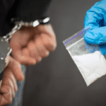 A police glove holding a baggie of white powder next to a person's hands in handcuffs, illustrating evidence subject to the exclusionary rule drug case Colorado.