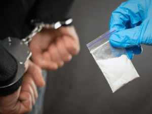 A police glove holding a baggie of white powder next to a person's hands in handcuffs, illustrating evidence subject to the exclusionary rule drug case Colorado.